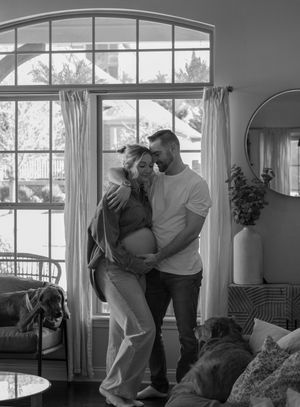 Black and white photo of smiling pregnant couple embracing in a bright living room with two dogs.