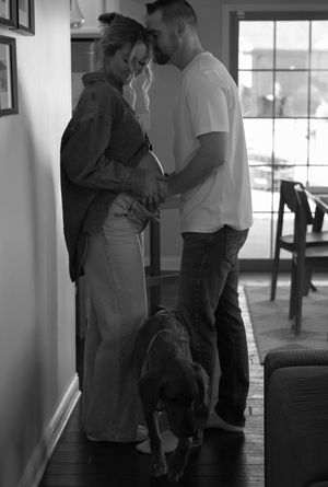 Black and white photo of expecting couple with dog looking at pregnant belly indoors.