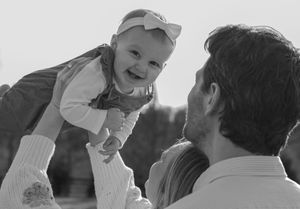 Black and white photo of happy baby being lifted up by parents outdoors in natural light.