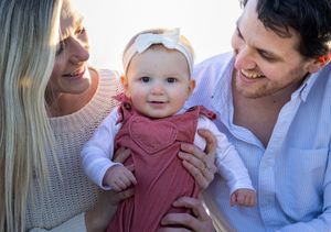 Smiling parents holding adorable baby girl in pink dress with white bow headband outdoors.