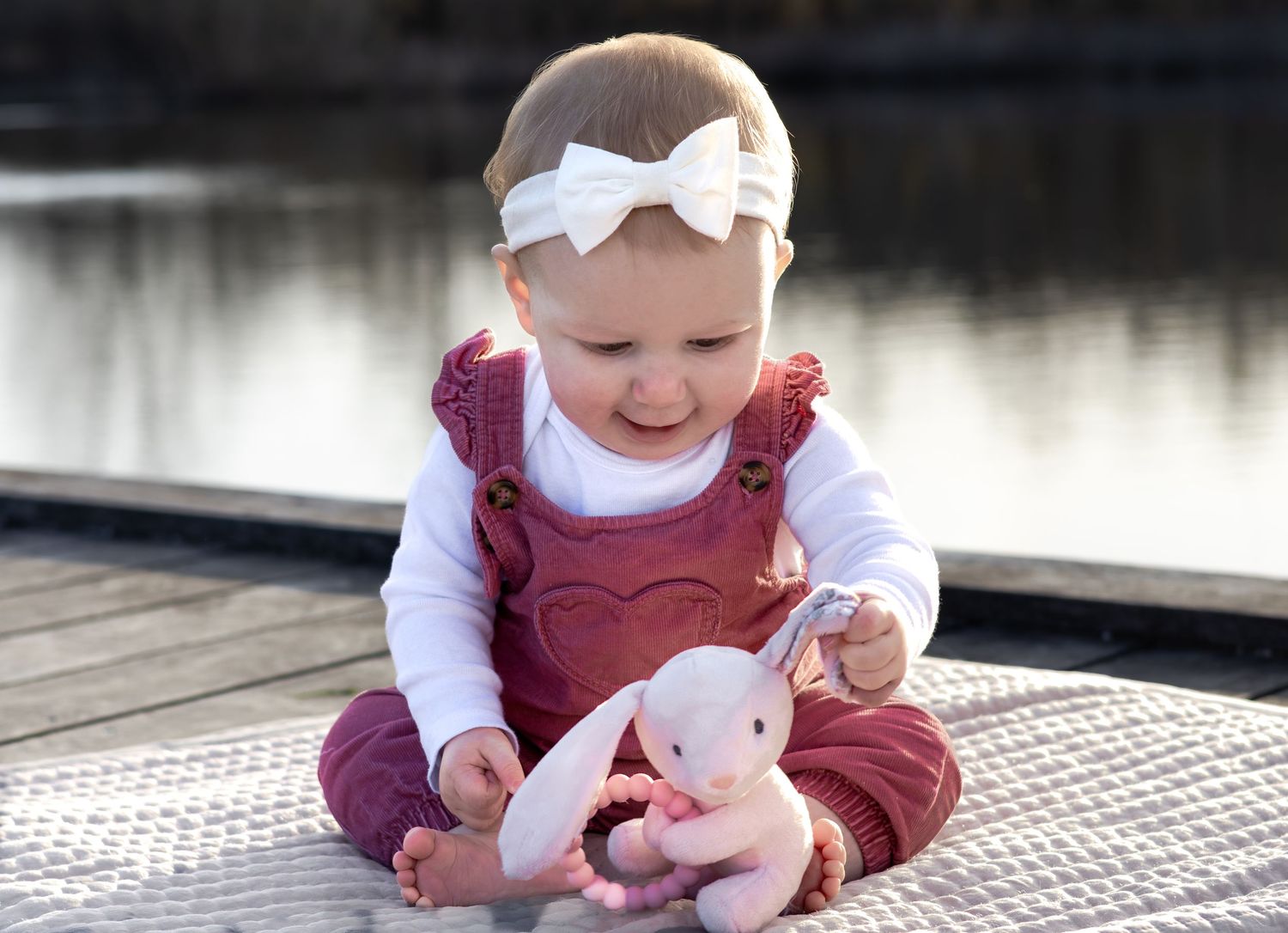 Smiling baby girl in red overalls and white bow headband playing with a pink bunny toy on a dock.