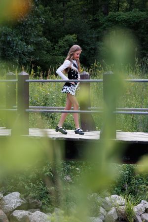A young girl in a black and white outfit walks across a wooden bridge surrounded by lush green trees and plants.