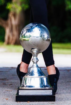 Person standing behind a large silver trophy with a globe on top, displayed outdoors on a paved surface.