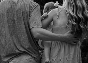 Black and white photo of a couple holding a baby girl with a bow, viewed from behind outdoors.