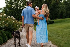Family walking on gravel path with baby and dog, surrounded by lush greenery, woman in blue dress, man in casual attire.