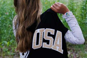 Girl wearing a black USA varsity jacket over a white lace long sleeve shirt, standing outdoors.