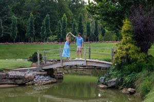 Couple in matching blue outfits dancing on a wooden bridge over a tranquil pond in a lush green garden.