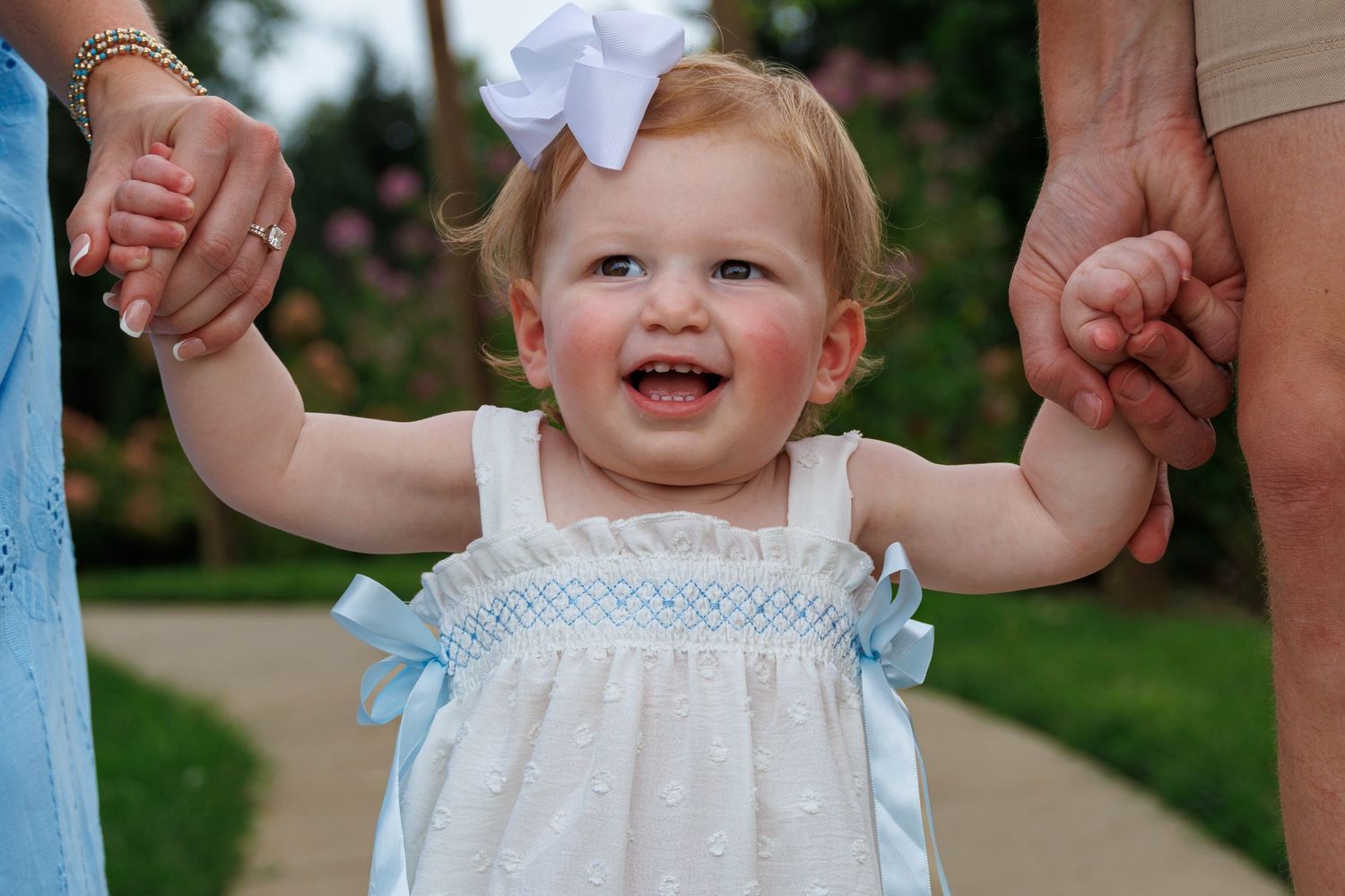 Smiling baby girl in white dress with blue bows and white hair bow, held up by adults outdoors.