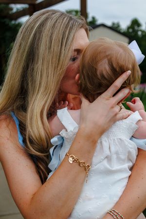 A blonde mother tenderly kisses her baby girl wearing a white dress and bow outdoors.