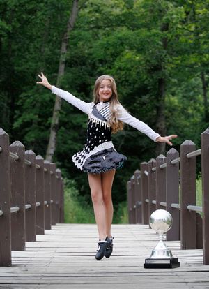 Young Irish dancer in black and white costume leaps joyfully on wooden bridge with trophy beside her.