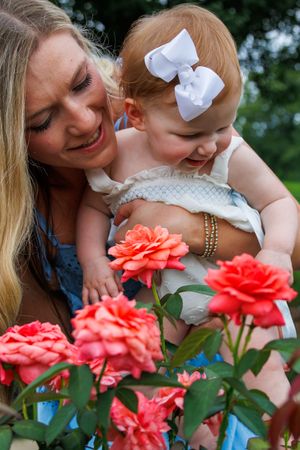 Mother and baby girl with white bow admiring pink roses in a garden, sharing a joyful outdoor moment.