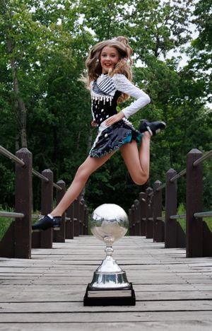 Young Irish dancer in black costume leaps over silver trophy on wooden bridge surrounded by green trees.