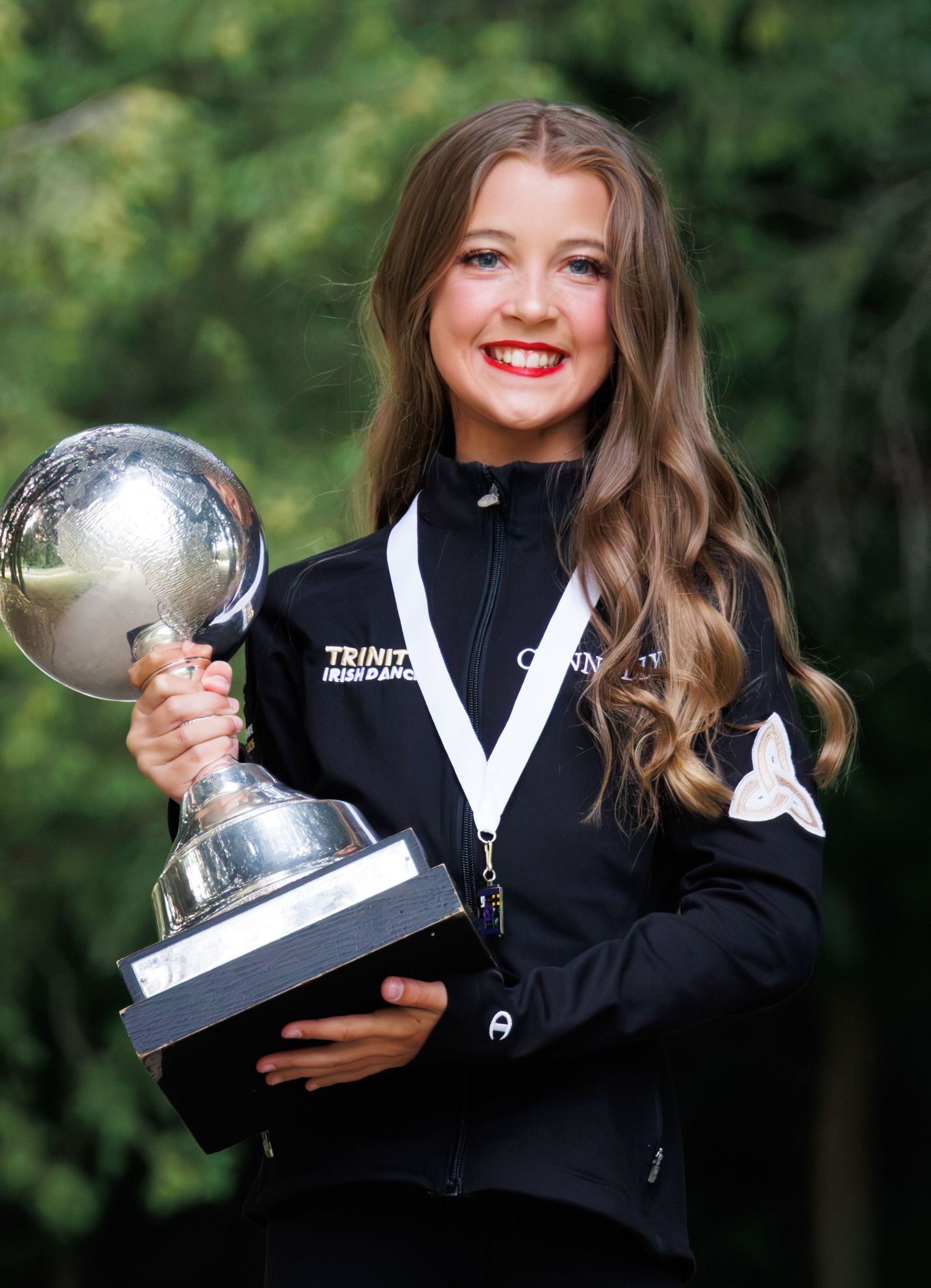 Young girl smiling proudly while holding a large trophy and wearing a medal and black jacket outdoors.