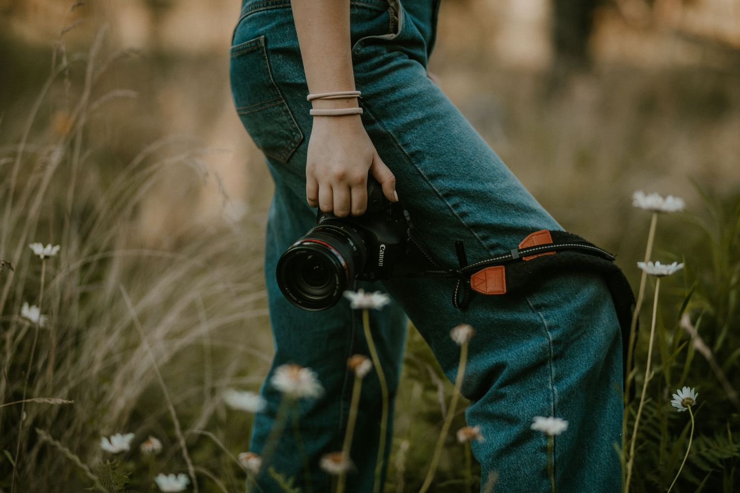 Person in teal overalls holding a DSLR camera while standing in a wildflower field with daisies.