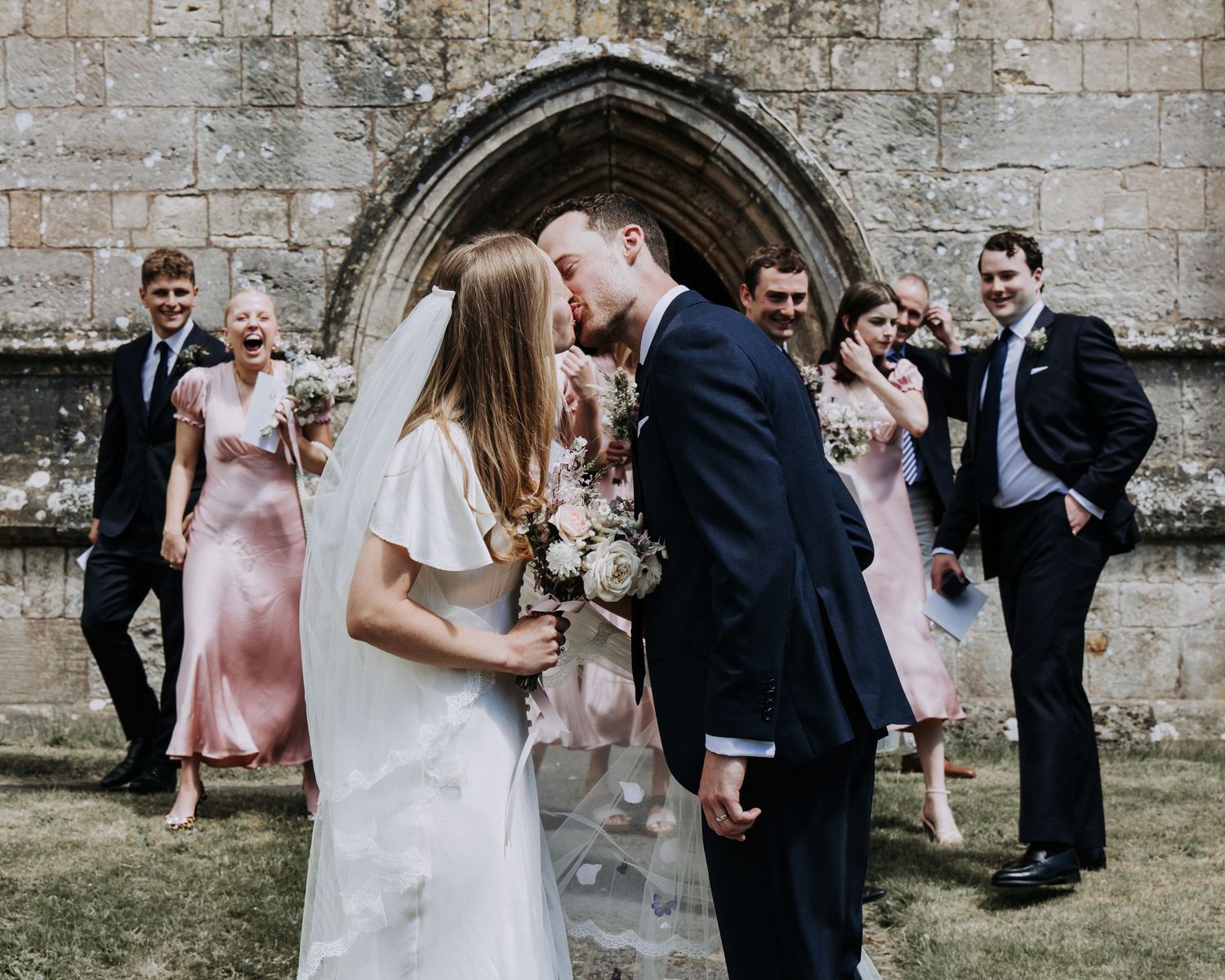 Newly married couple sharing their first kiss outside the church surrounded by their bridal party.