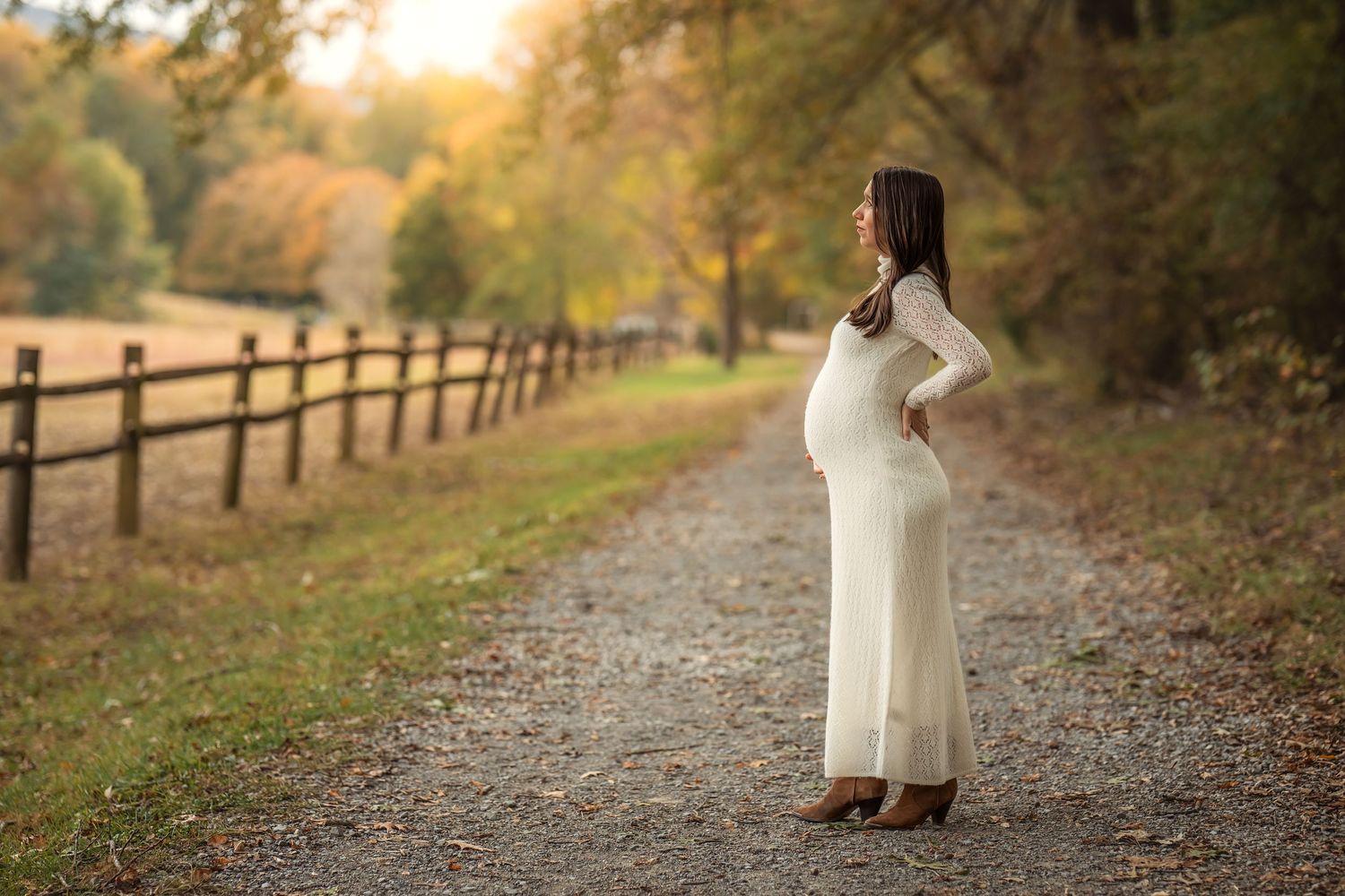 Pregnant woman in white dress standing on autumn path by wooden fence.