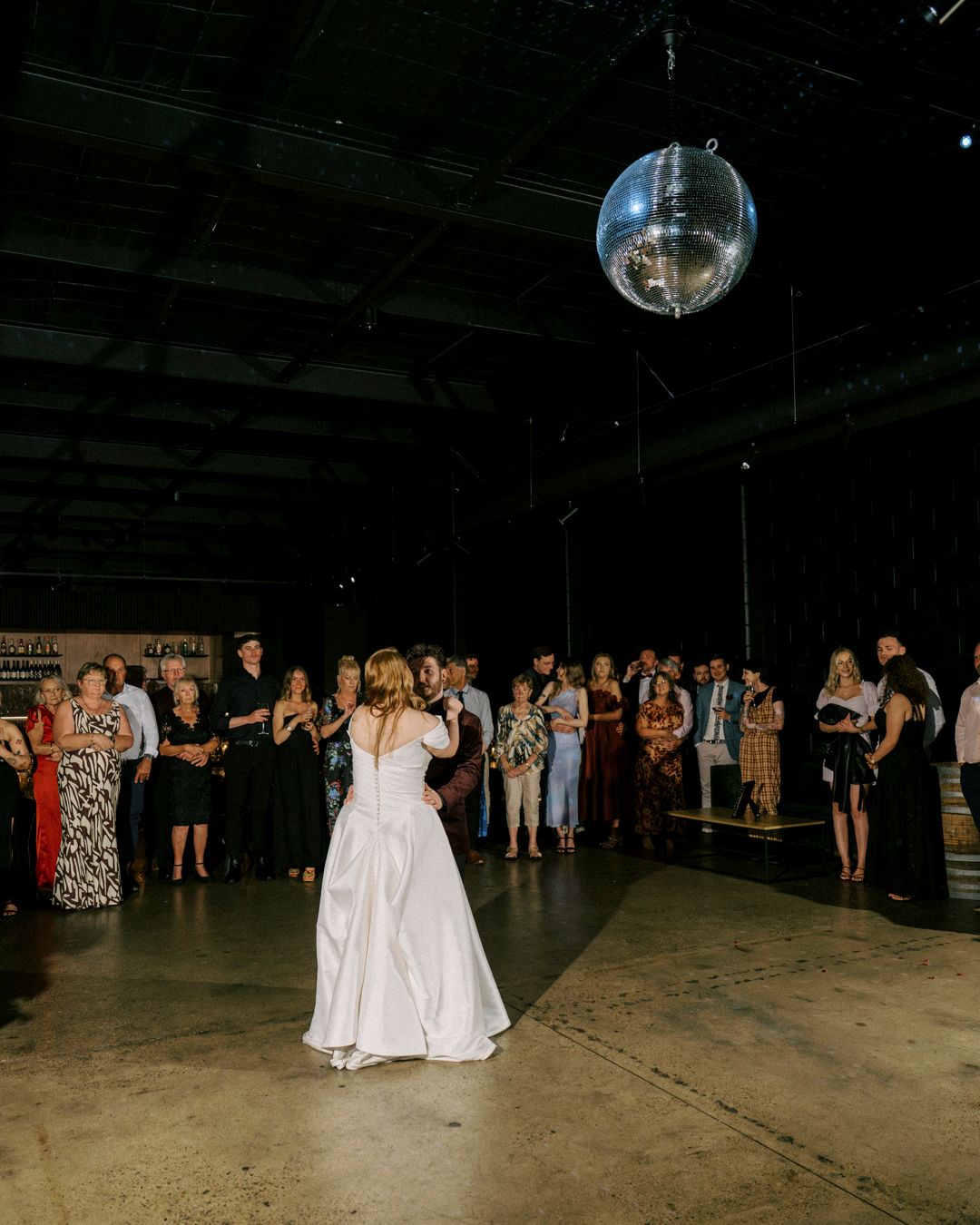 Bride in white gown stands under disco ball at industrial wedding venue, guests watching in background.the bethnal. Newstead Brisbane