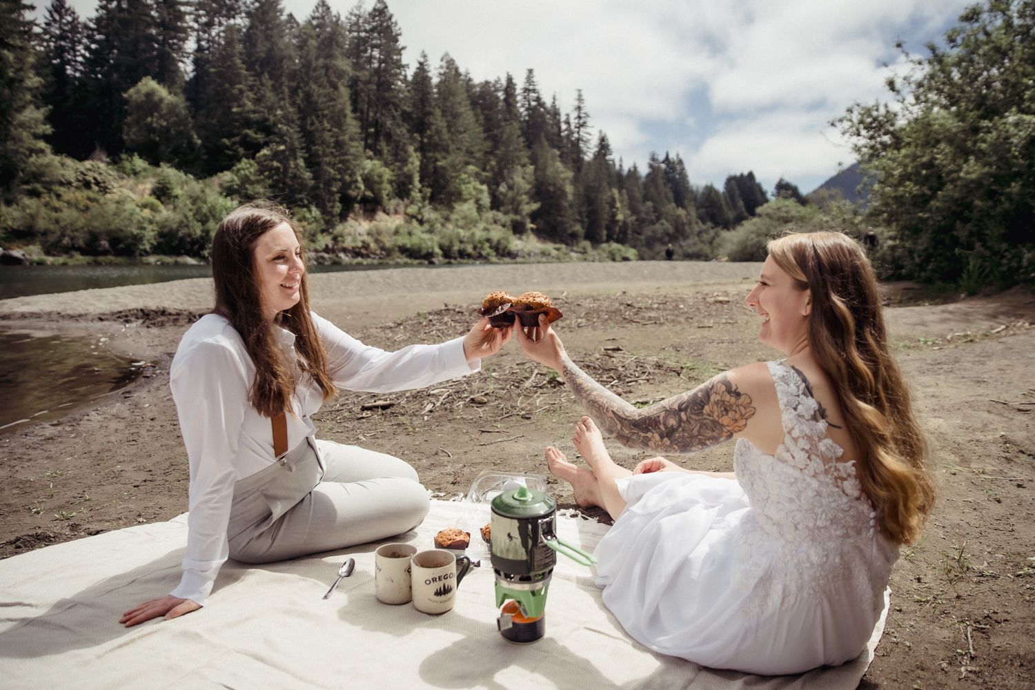 Two brides share muffins during an elopement day brunch picnic, with coffee and a picnic blanket by the smith river
