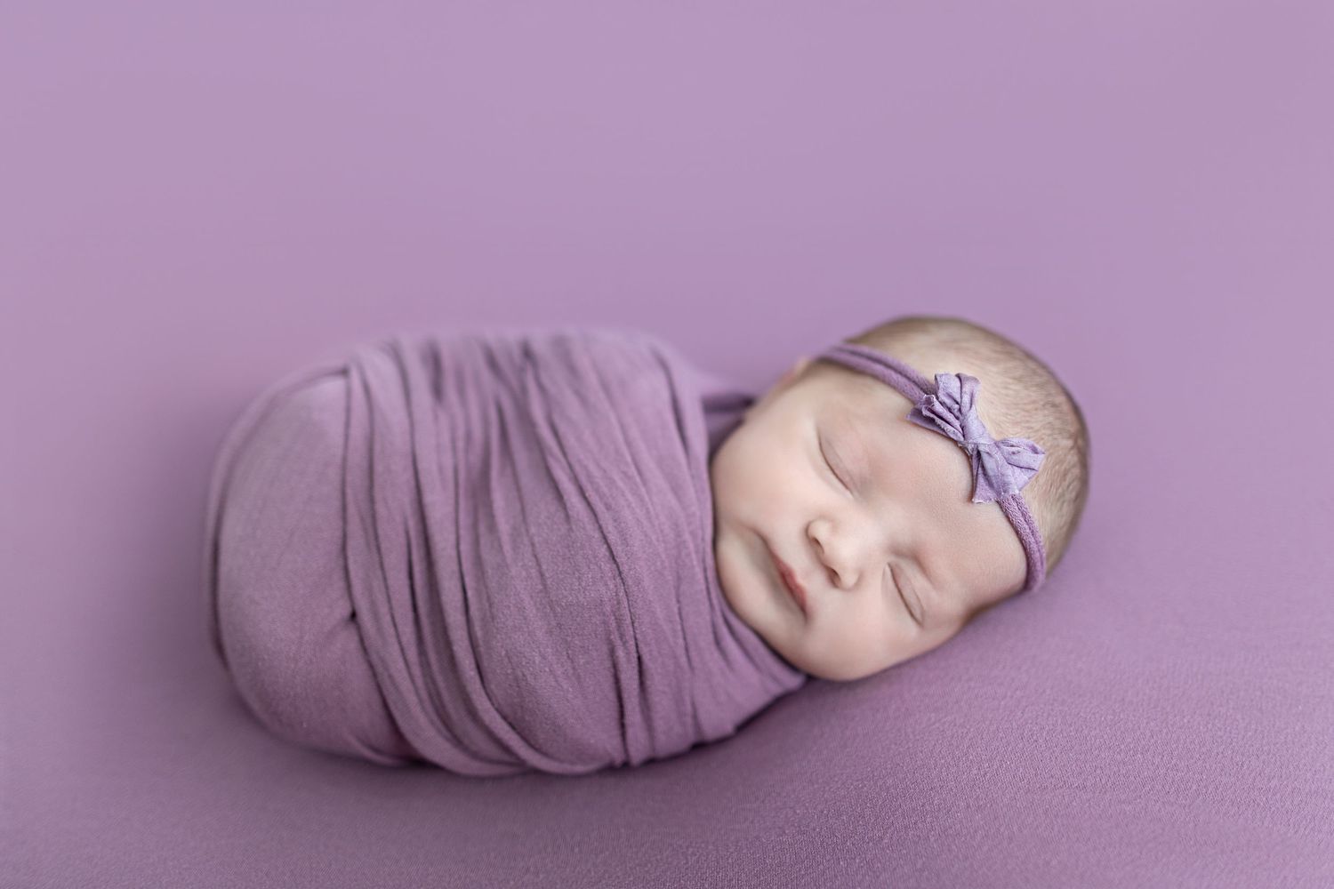 Sleeping newborn baby girl swaddled in purple wrap with bow headband on matching lavender background.