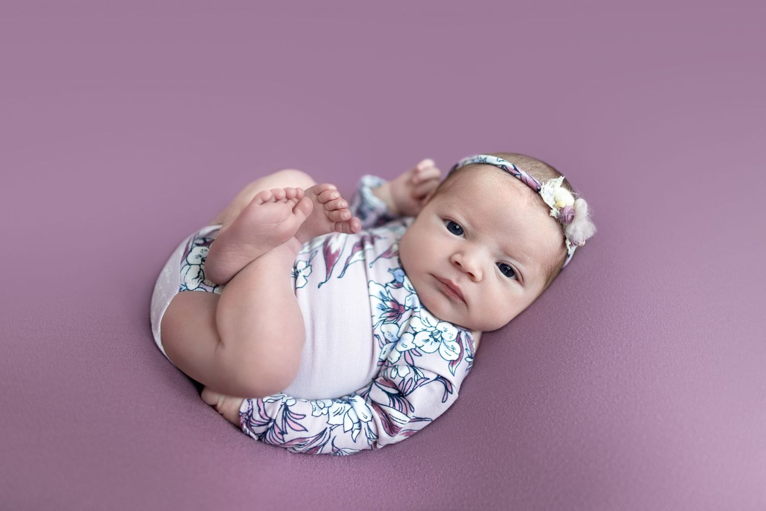 Newborn baby girl in floral romper and bow headband posed on purple backdrop for professional photo.