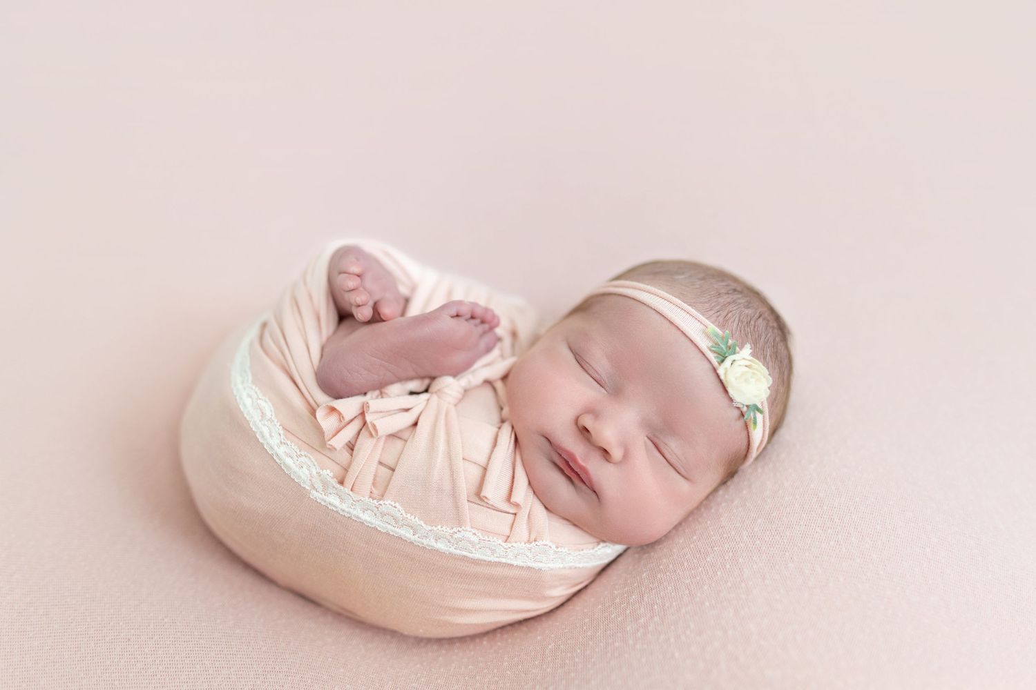 Newborn baby girl swaddled in pink wrap sleeping peacefully on a soft pink backdrop with a delicate floral headband.