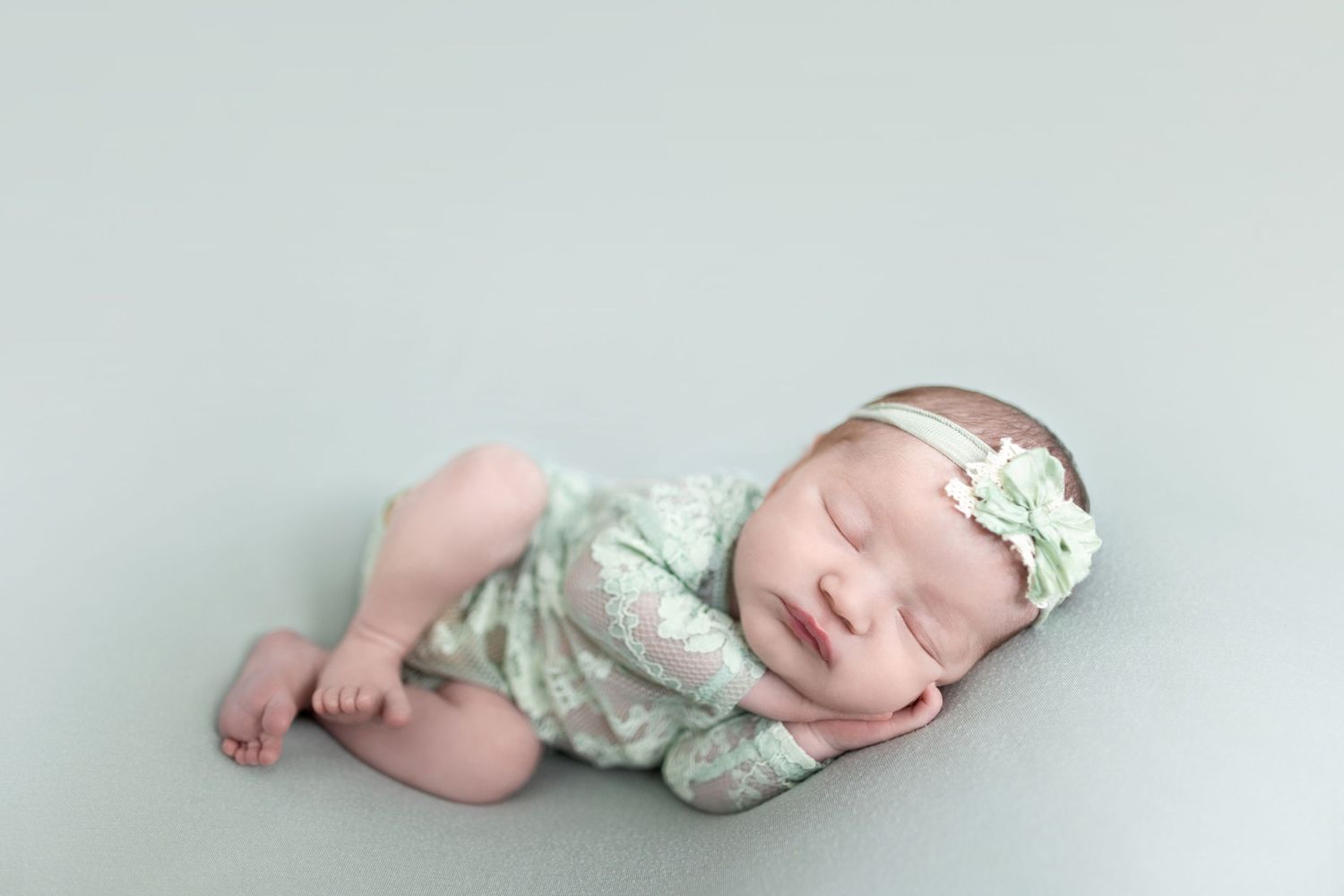 Sleeping newborn baby girl wrapped in mint green swaddle with floral headband on soft teal background.