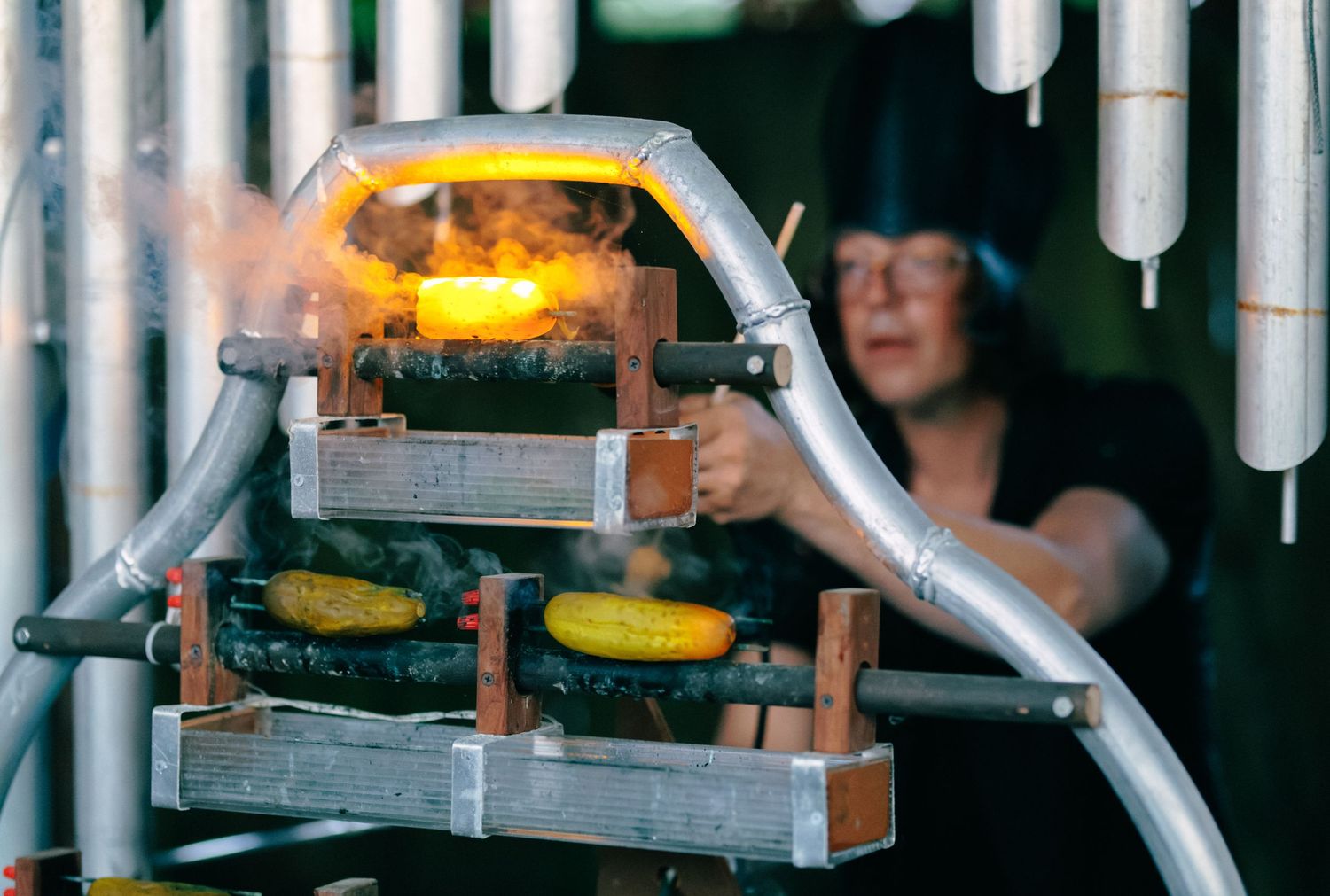 Glassblower using a furnace to heat molten glass, with flames glowing orange in an outdoor workshop setting.