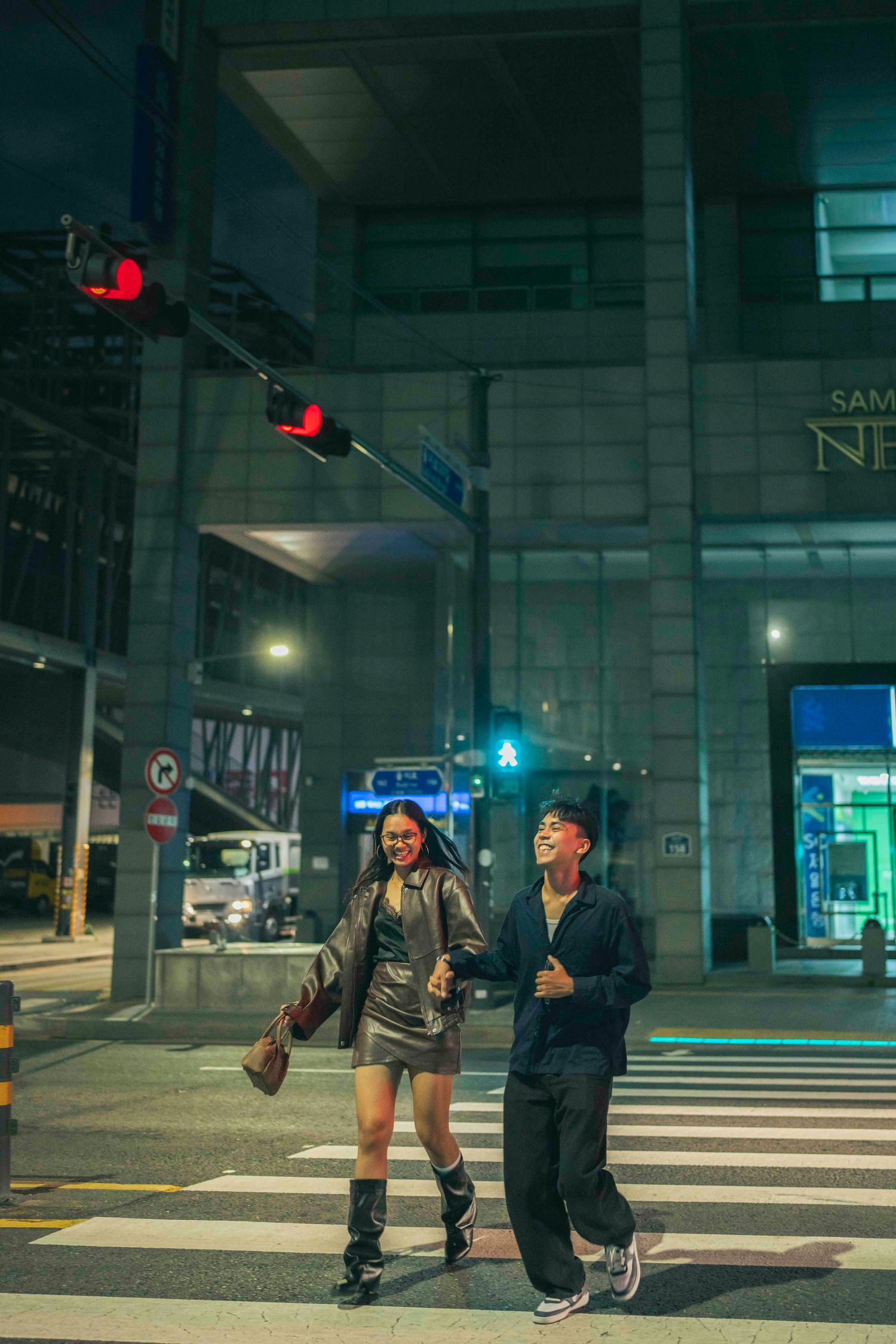 Two young people laughing and crossing a city street at night near a modern building with red traffic lights.