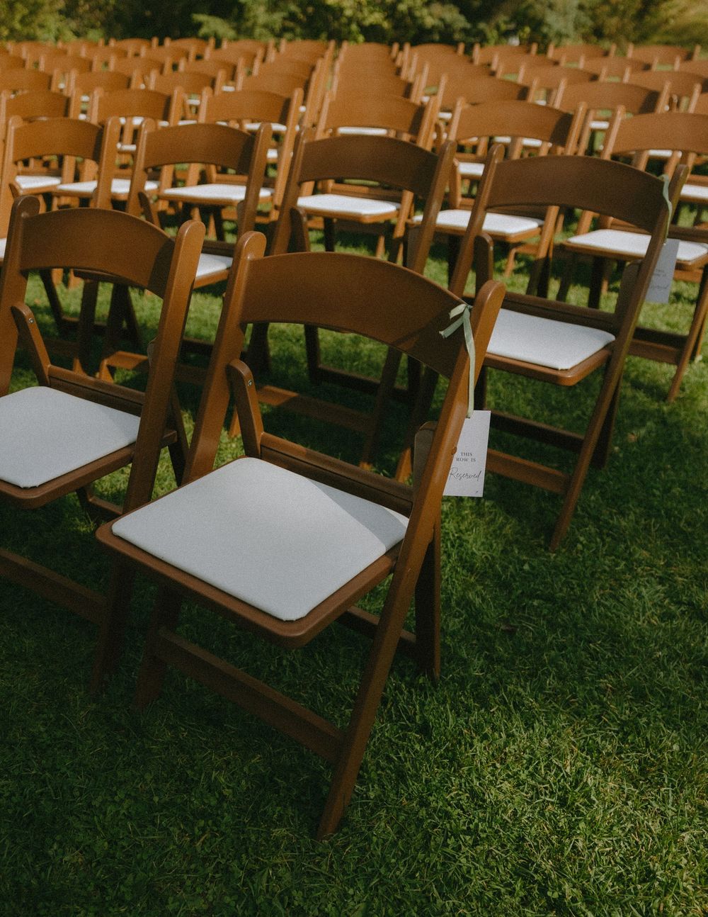 Rows of wooden folding chairs with white cushions arranged on green grass for an outdoor wedding ceremony.