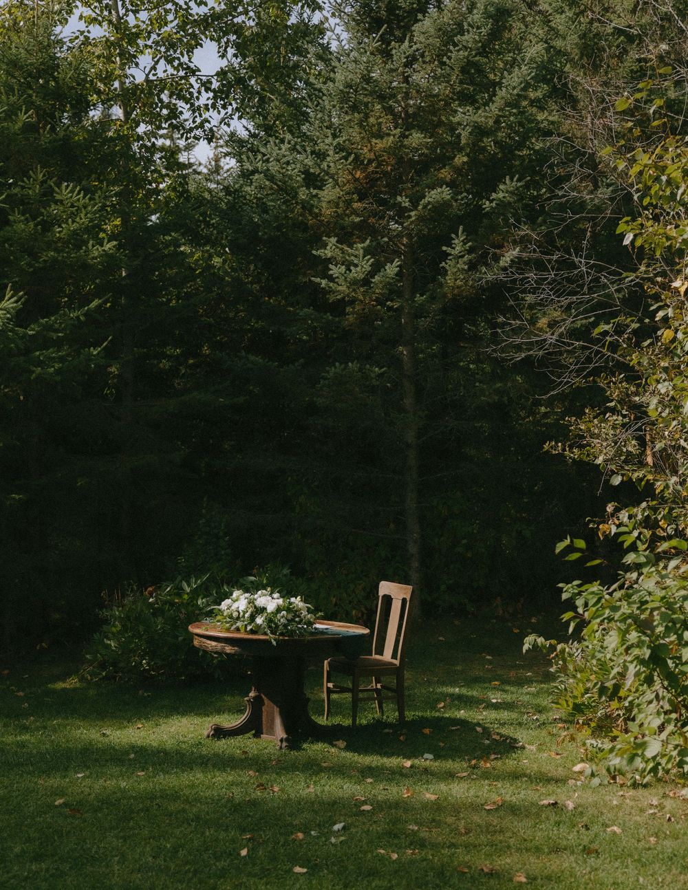 Rustic wooden table with white floral centerpiece beside a chair on a sunlit lawn surrounded by lush green forest.