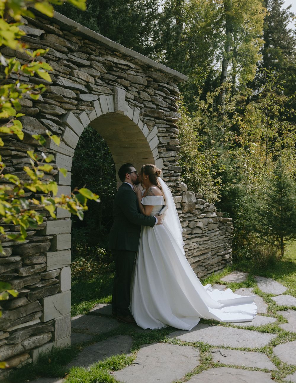 Bride and groom sharing a romantic moment under a rustic stone arch surrounded by lush greenery.