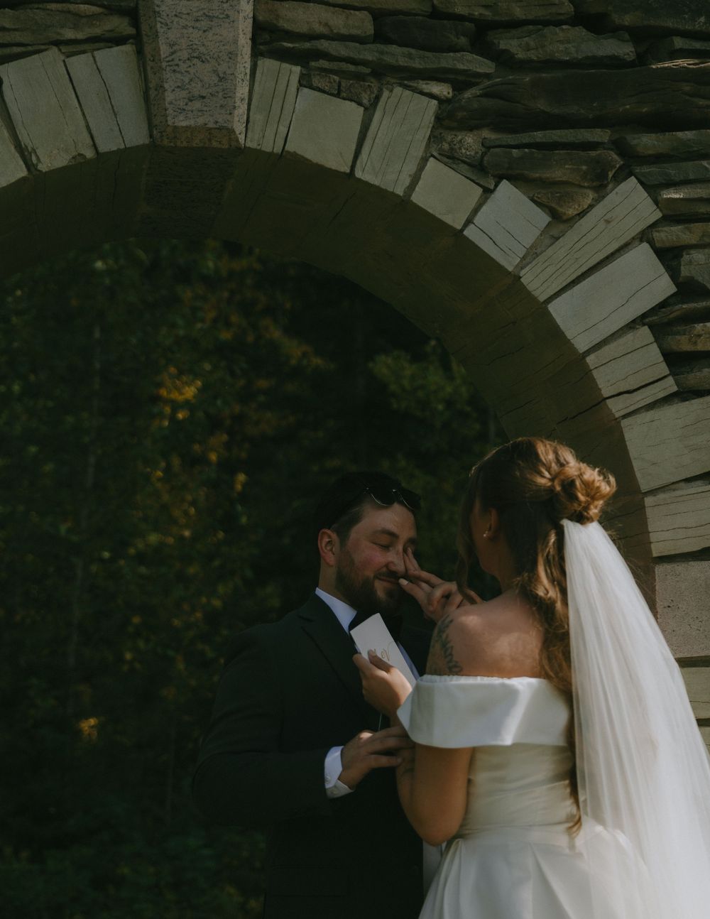 Bride and groom share an intimate moment under a stone arch, bride in strapless gown with veil, groom in black tuxedo.