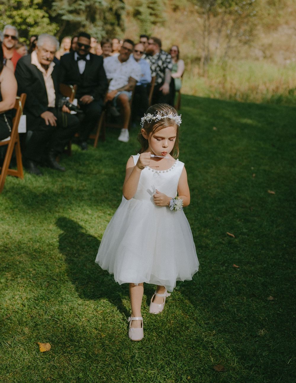 A flower girl in a white tulle dress walks down a grassy outdoor wedding aisle lined with seated guests.