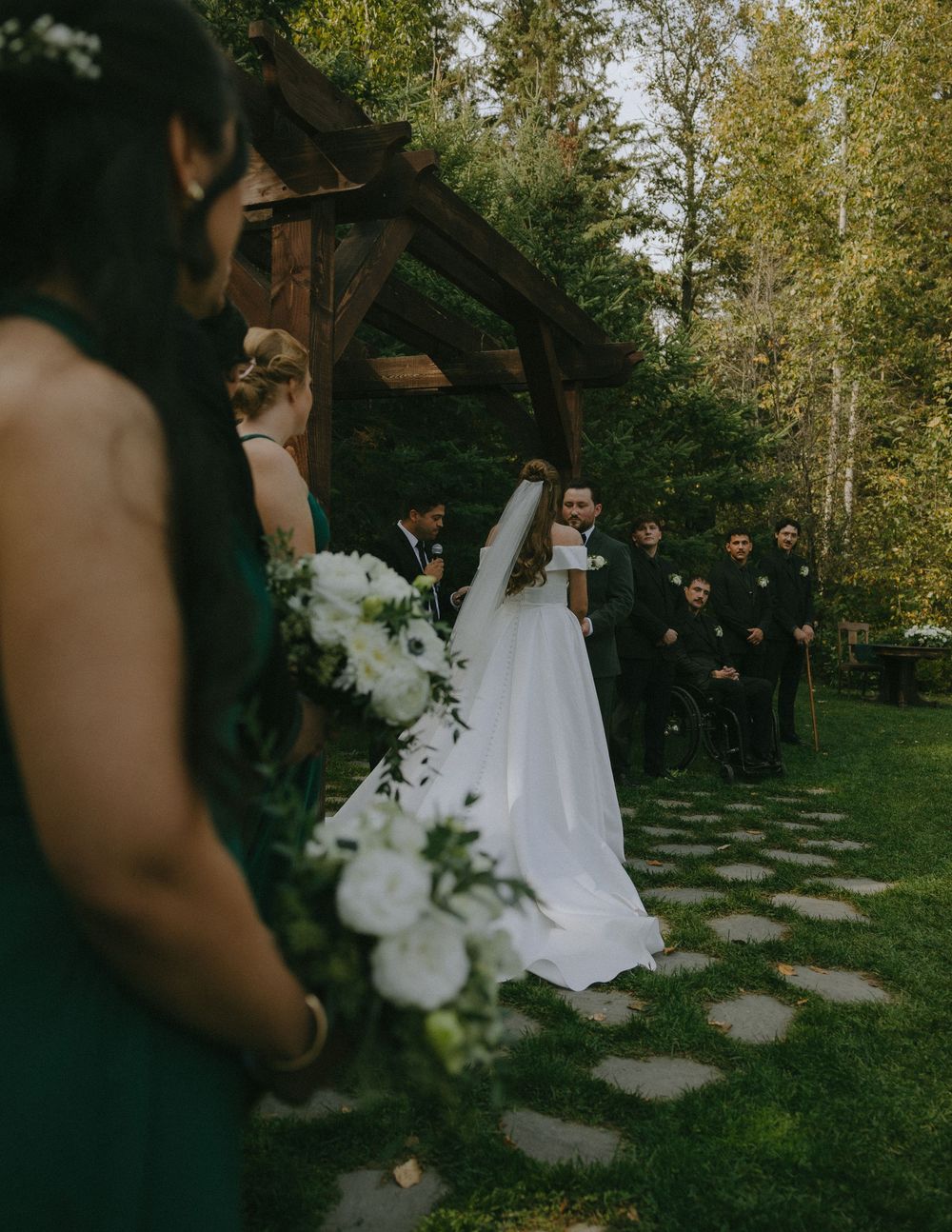 Outdoor wedding ceremony under a wooden arbor with bridesmaids in emerald green dresses and guests on a stone path.