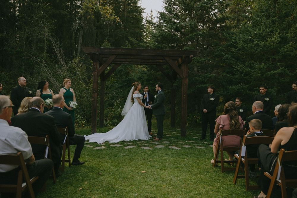 Bride and groom exchange vows at an outdoor woodland wedding ceremony under a rustic wooden arch.