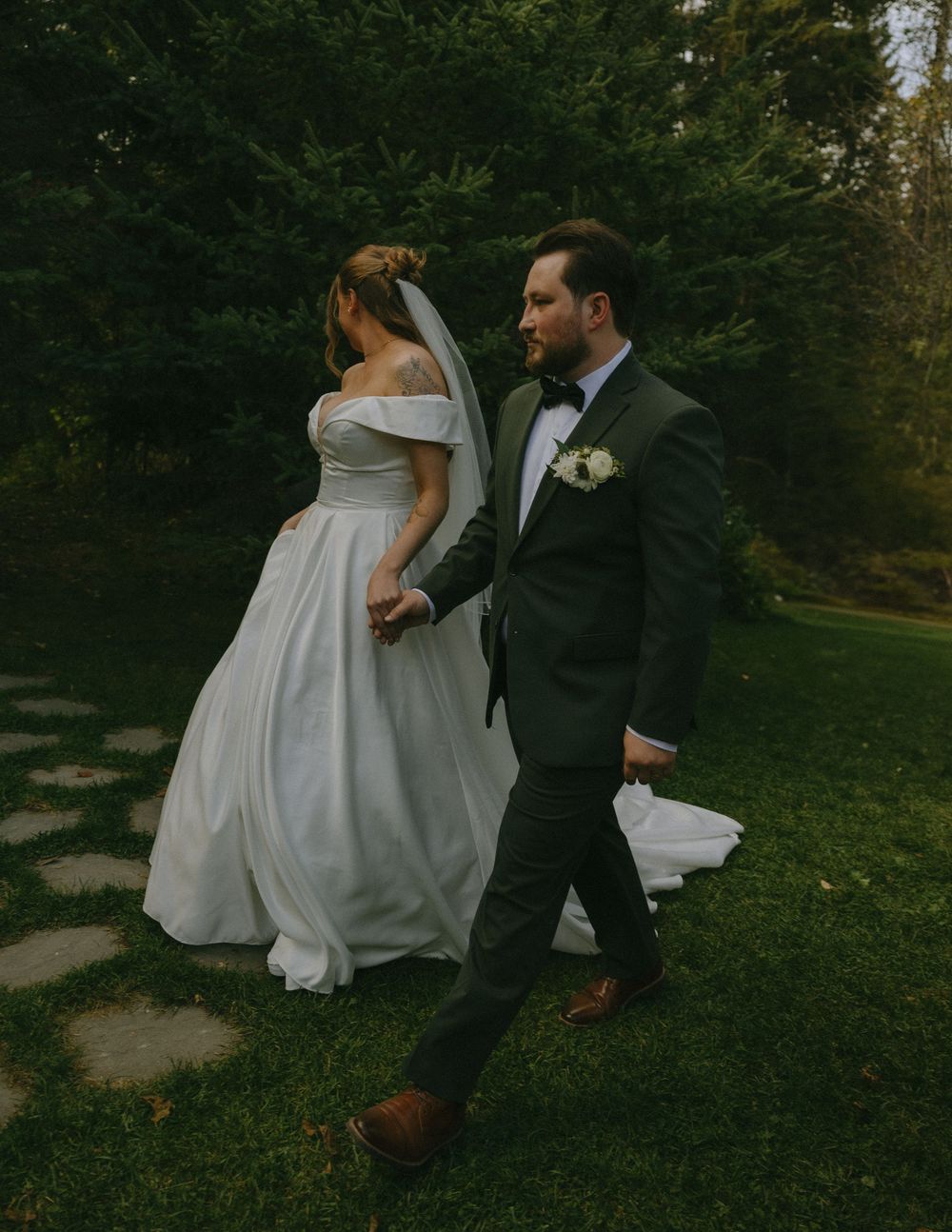 Bride and groom walking hand in hand through lush green garden in elegant wedding attire.