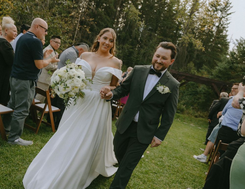 Happy newlyweds walk down outdoor wedding aisle, bride in white gown with bouquet, groom in dark tuxedo.