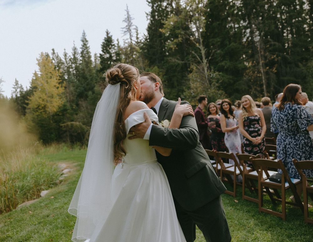 Bride and groom share first kiss at outdoor forest wedding ceremony with guests seated in wooden chairs.