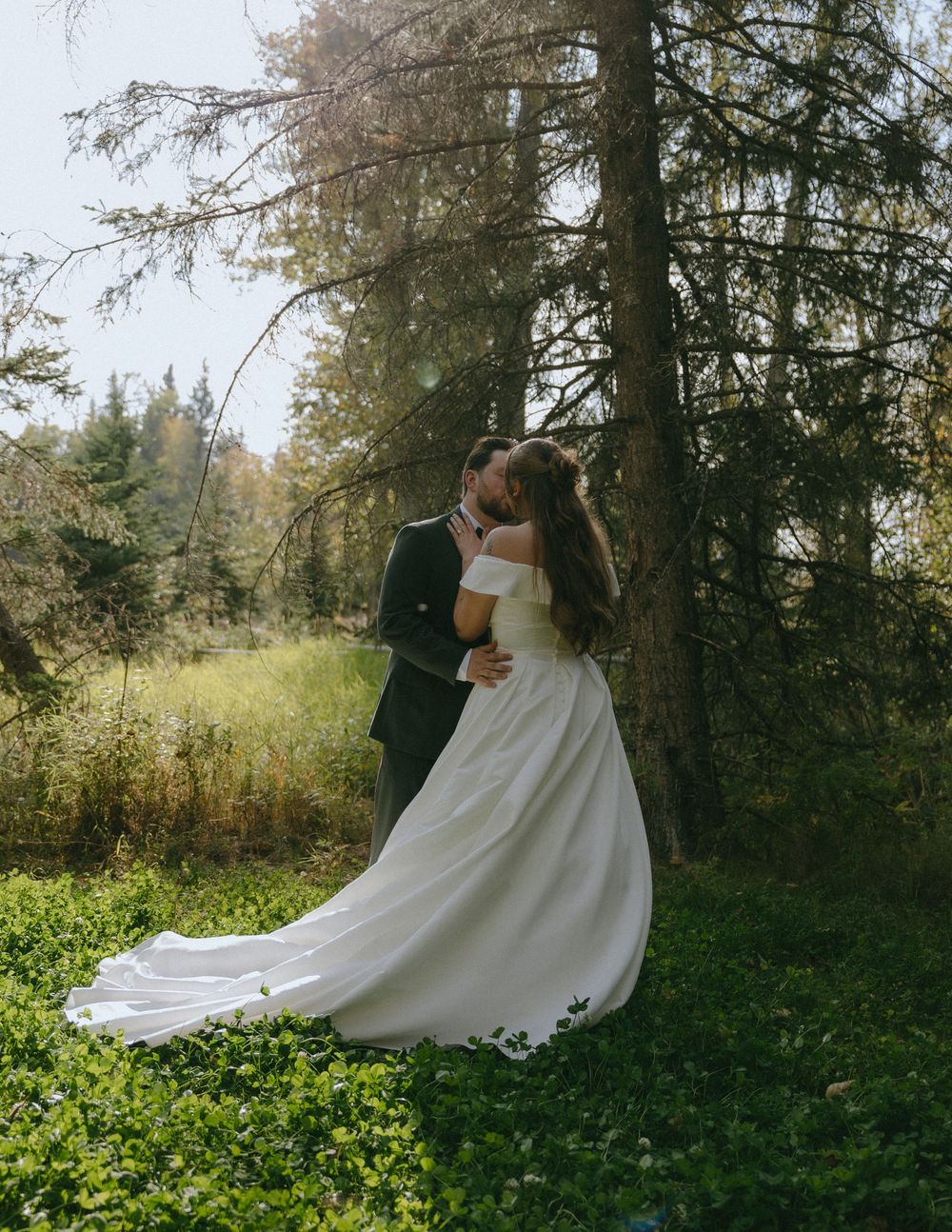 A bride and groom share a romantic kiss in a lush forest setting, her white gown trailing on the green ground.