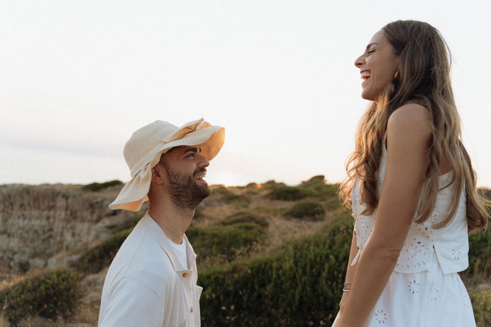 A smiling couple in white outfits enjoys a sunset outdoors, man wearing a straw hat, woman with long wavy hair.