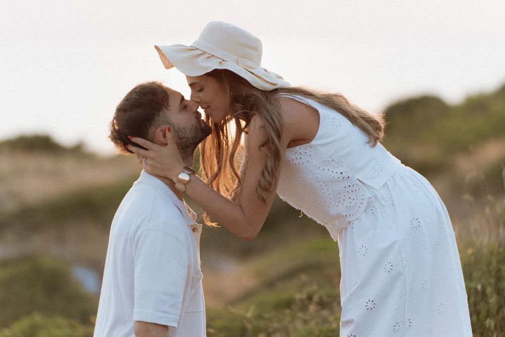 Romantic couple in white outfits sharing a kiss outdoors at sunset, woman wearing a wide-brim hat.