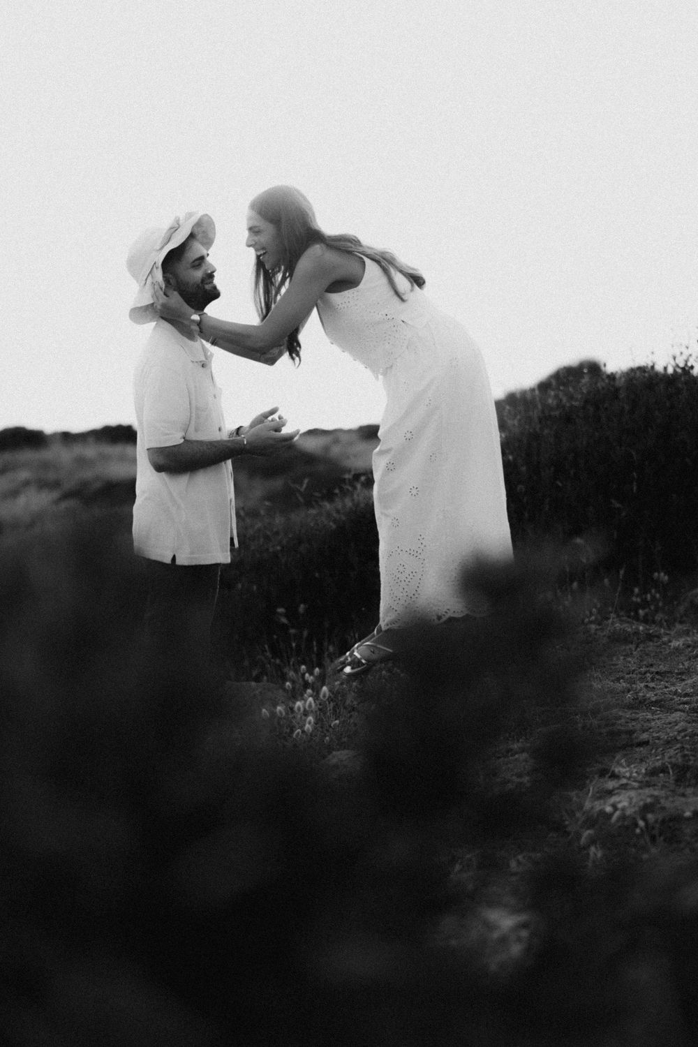 Black and white photo of a couple in white outfits sharing a tender moment in a field.