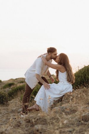Couple sharing a romantic kiss at sunset on a scenic hillside, woman in white dress seated among wildflowers.