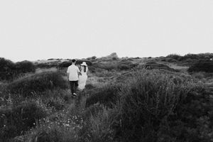 A couple walks hand-in-hand through a lush meadow landscape in a romantic black and white photo.