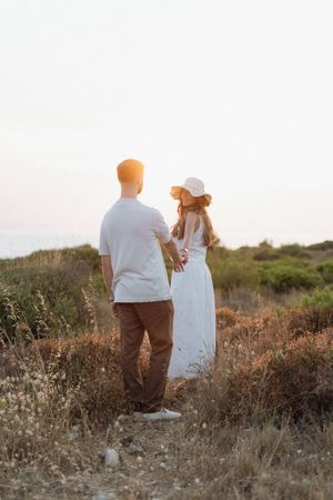 Couple holding hands in a golden field at sunset, woman in white dress and hat, man in white shirt and brown pants.