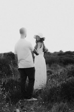 Black and white photo of a couple holding hands in a field, woman in white dress and sun hat, man in casual attire.
