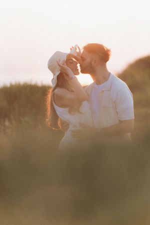 Couple sharing a romantic kiss at golden hour sunset in a field, woman wearing a white hat and dress.