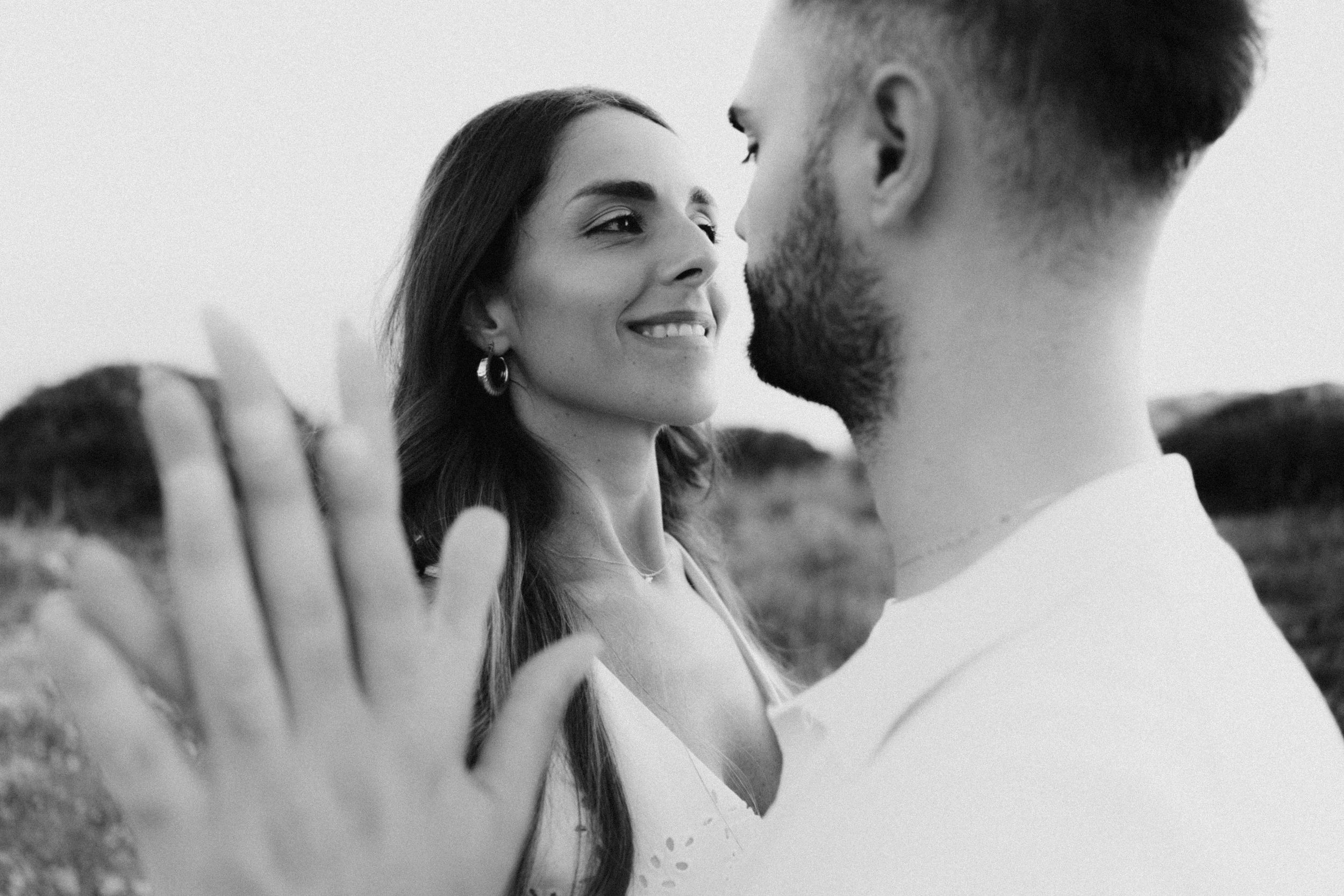 Black and white photo of a smiling woman and bearded man sharing a romantic moment outdoors.