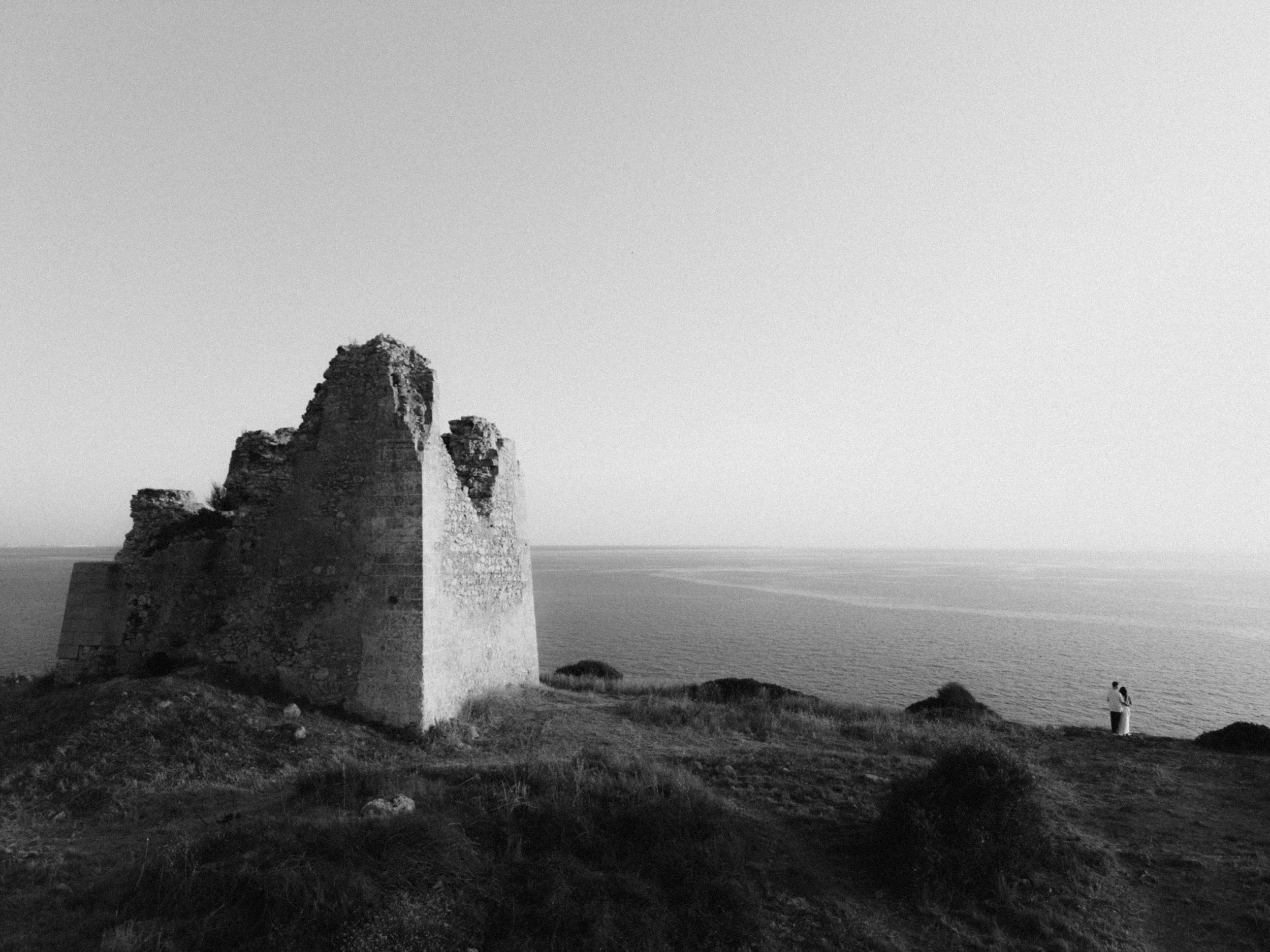 Black and white photo of ancient stone ruins on a coastal cliff overlooking a calm sea horizon.