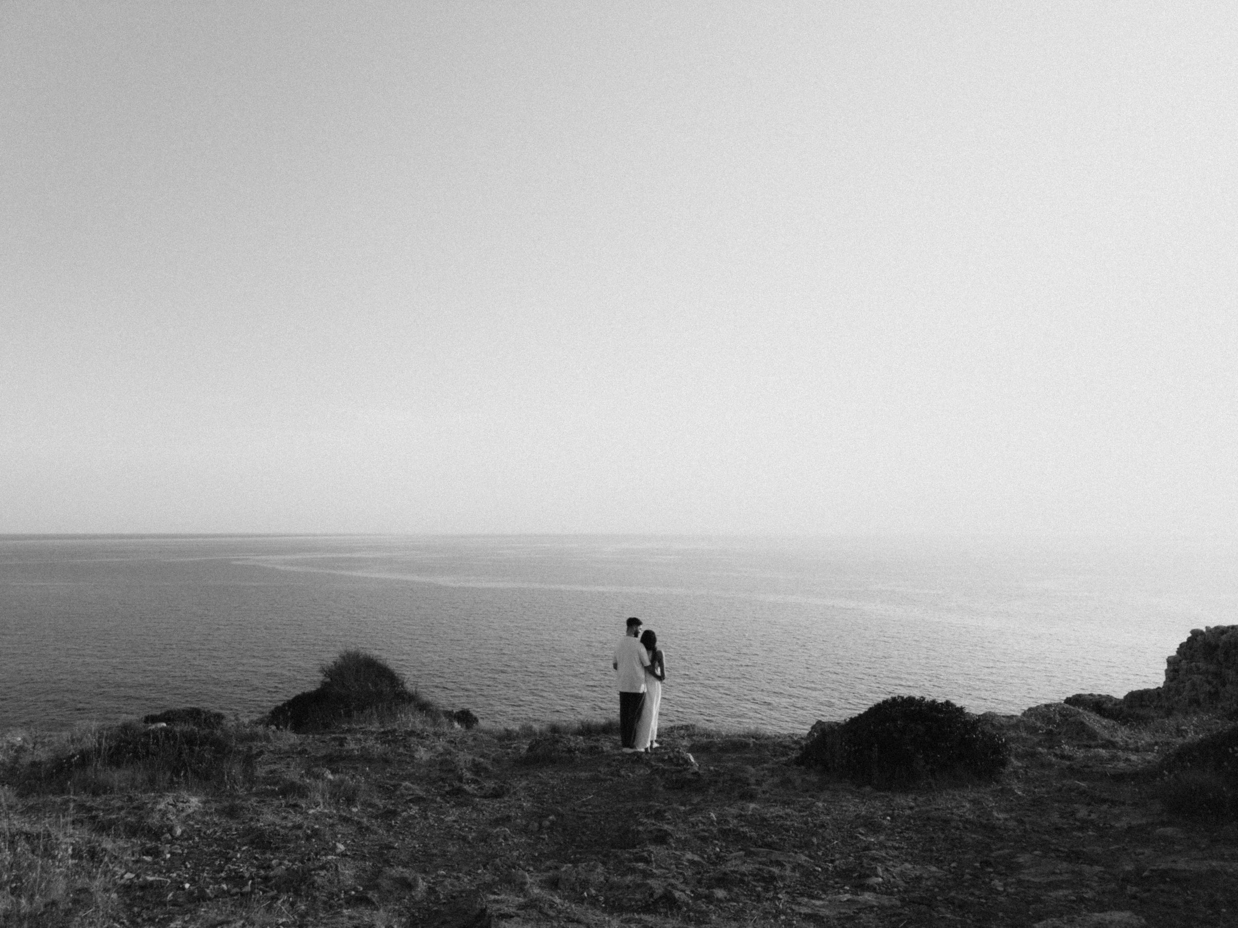 Black and white photo of a couple embracing on a rocky cliff overlooking a vast, calm ocean horizon.